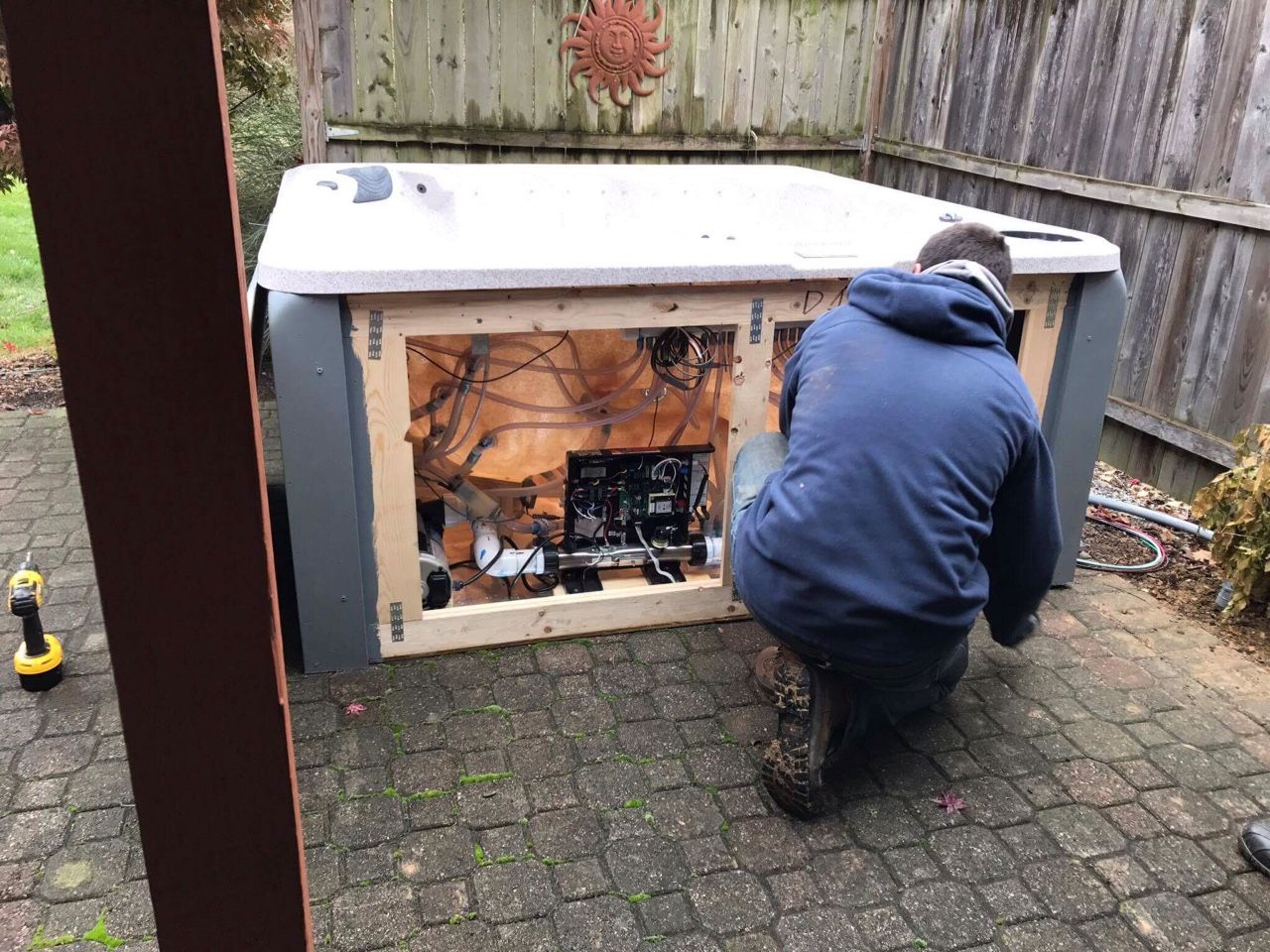 A man kneeling to repair the open side of a hot tub on a patio.