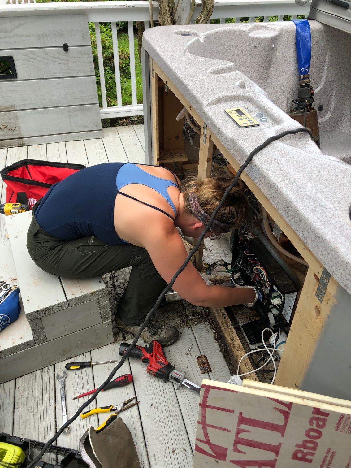 Woman fixing a hot tub on a wooden deck; she's in dark pants and a blue tank top, tools are scattered around.