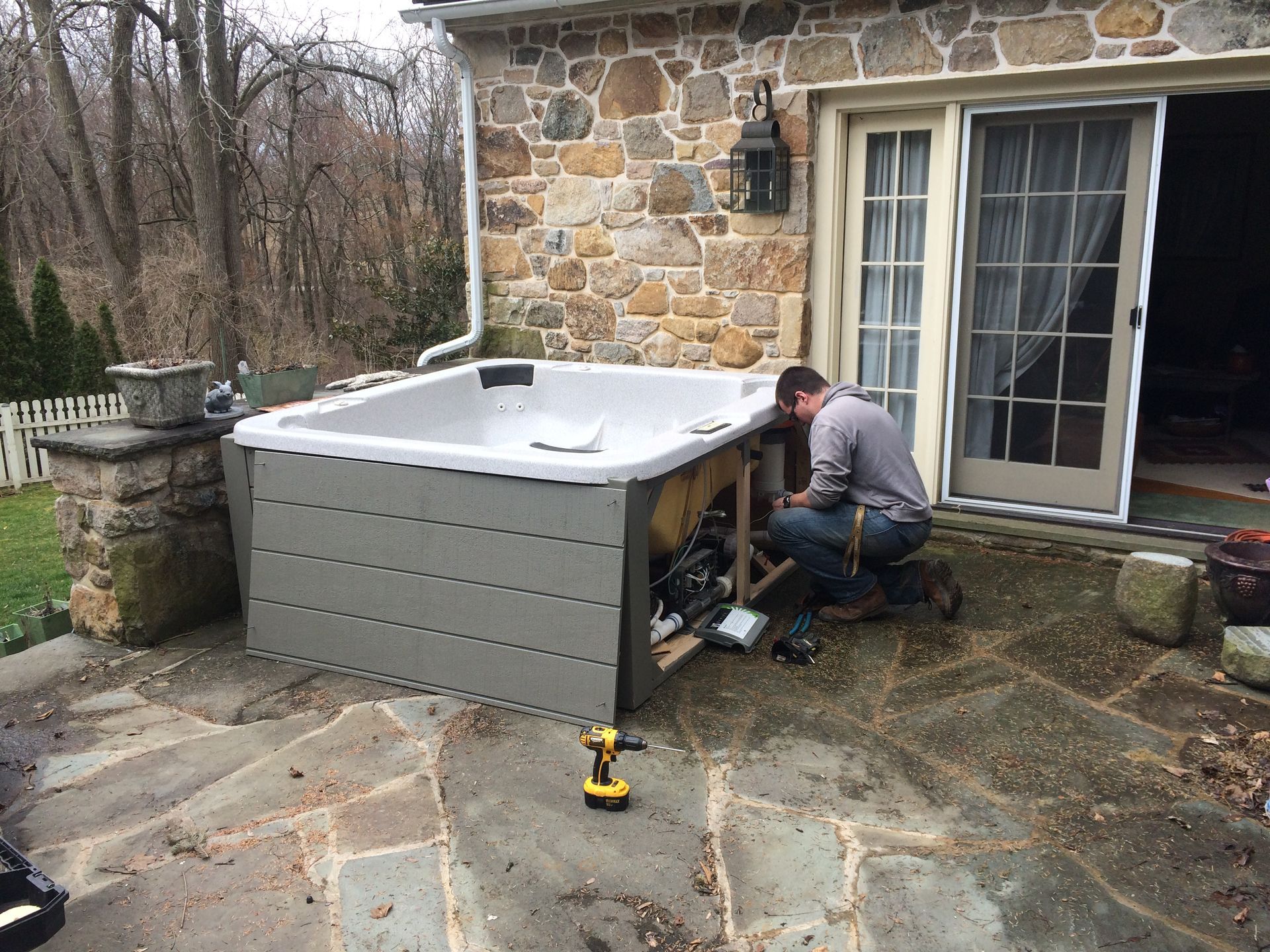 Man installing a hot tub on a stone patio next to a stone building with a sliding door.