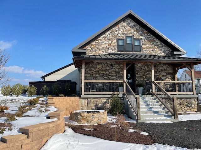 Stone-clad house with a porch, steps, and tiered landscaping in a snowy setting under a blue sky.