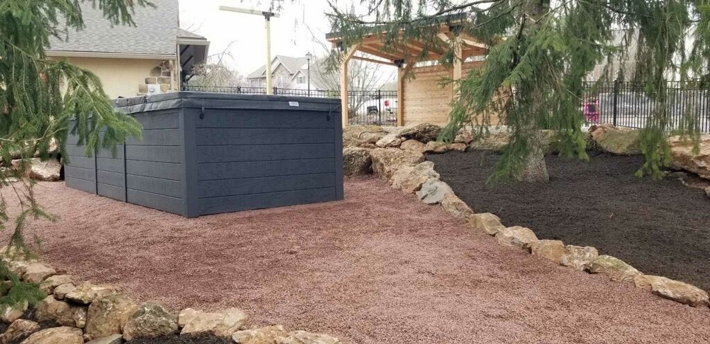Hot tub area in a yard; dark tub, red gravel, landscaping with a pergola and tree.