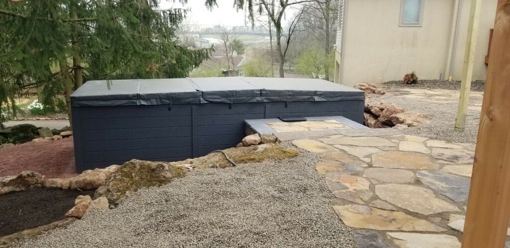 A covered, dark-colored hot tub sits on a stone patio surrounded by gravel and landscaping. A house is in the background.