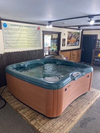 Hot tub with water sits on wooden pallet in a shop. Brown and green colors, overhead lights.