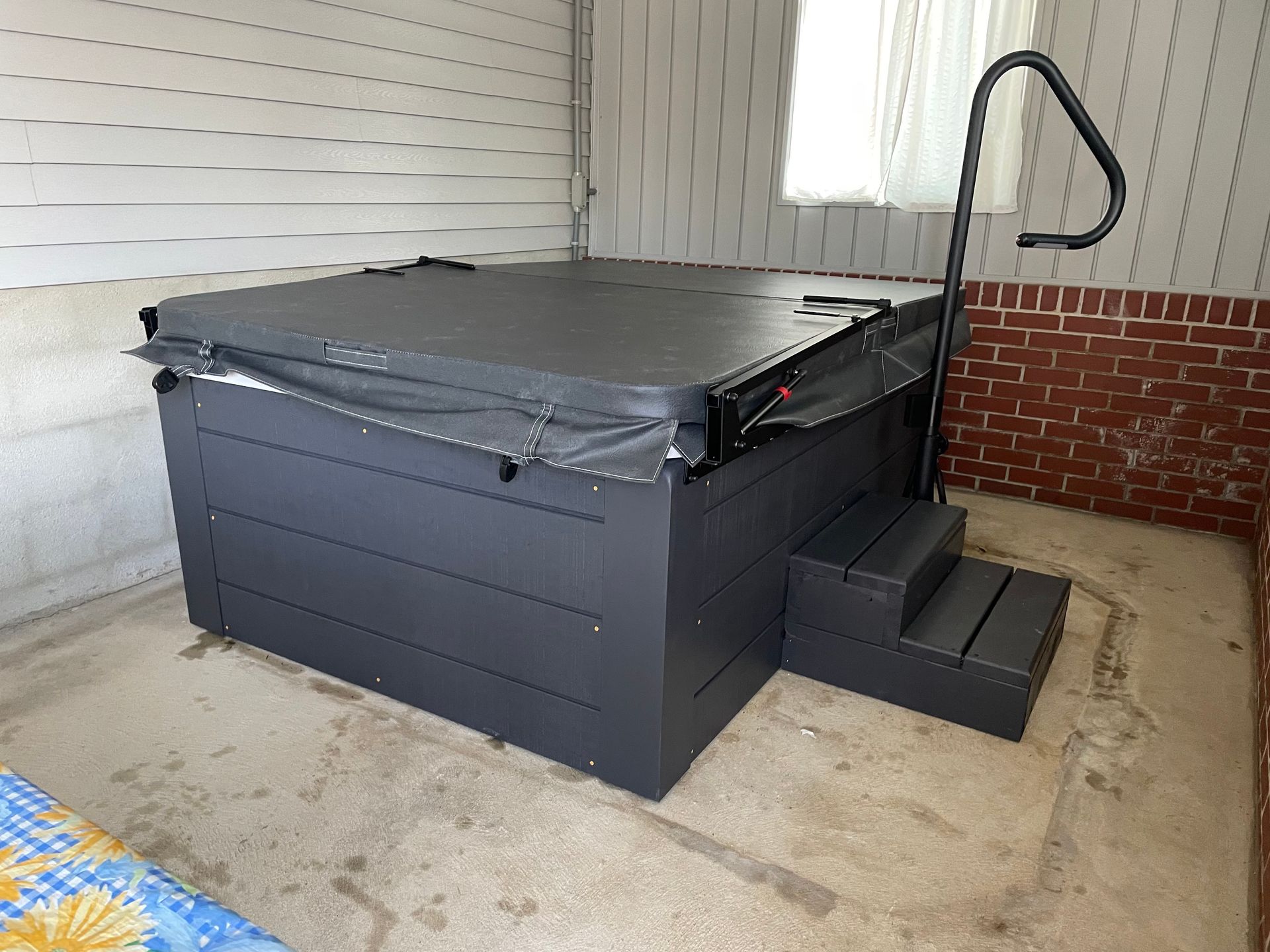 Long, rectangular swim spa under a metal shelter, filled with water; surrounded by grass and trees.