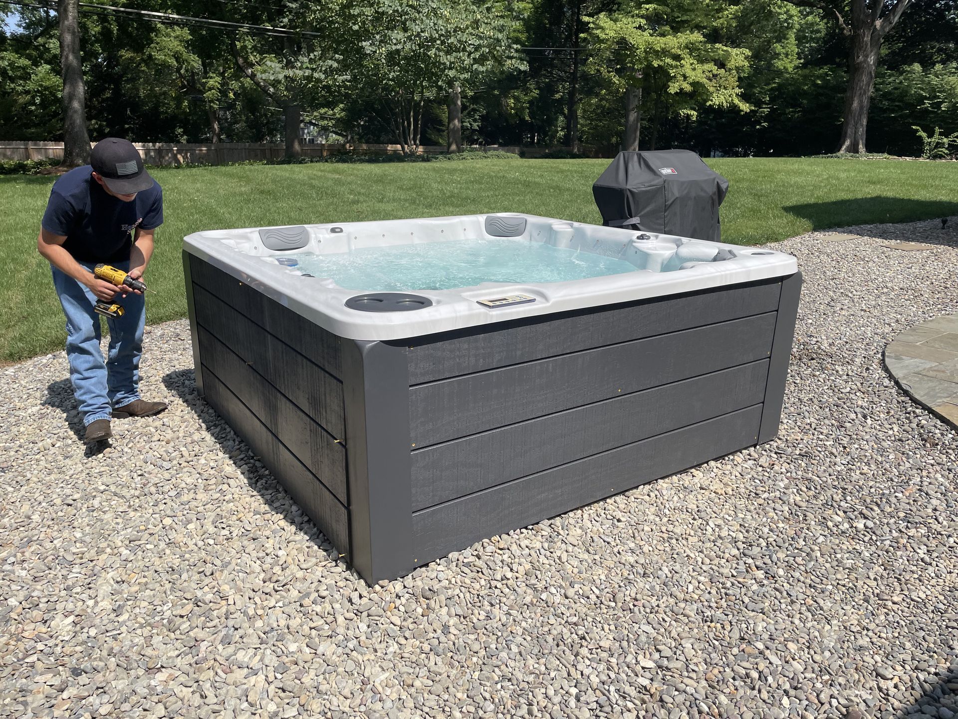 Man by a filled, gray hot tub on gravel patio; grill in background.
