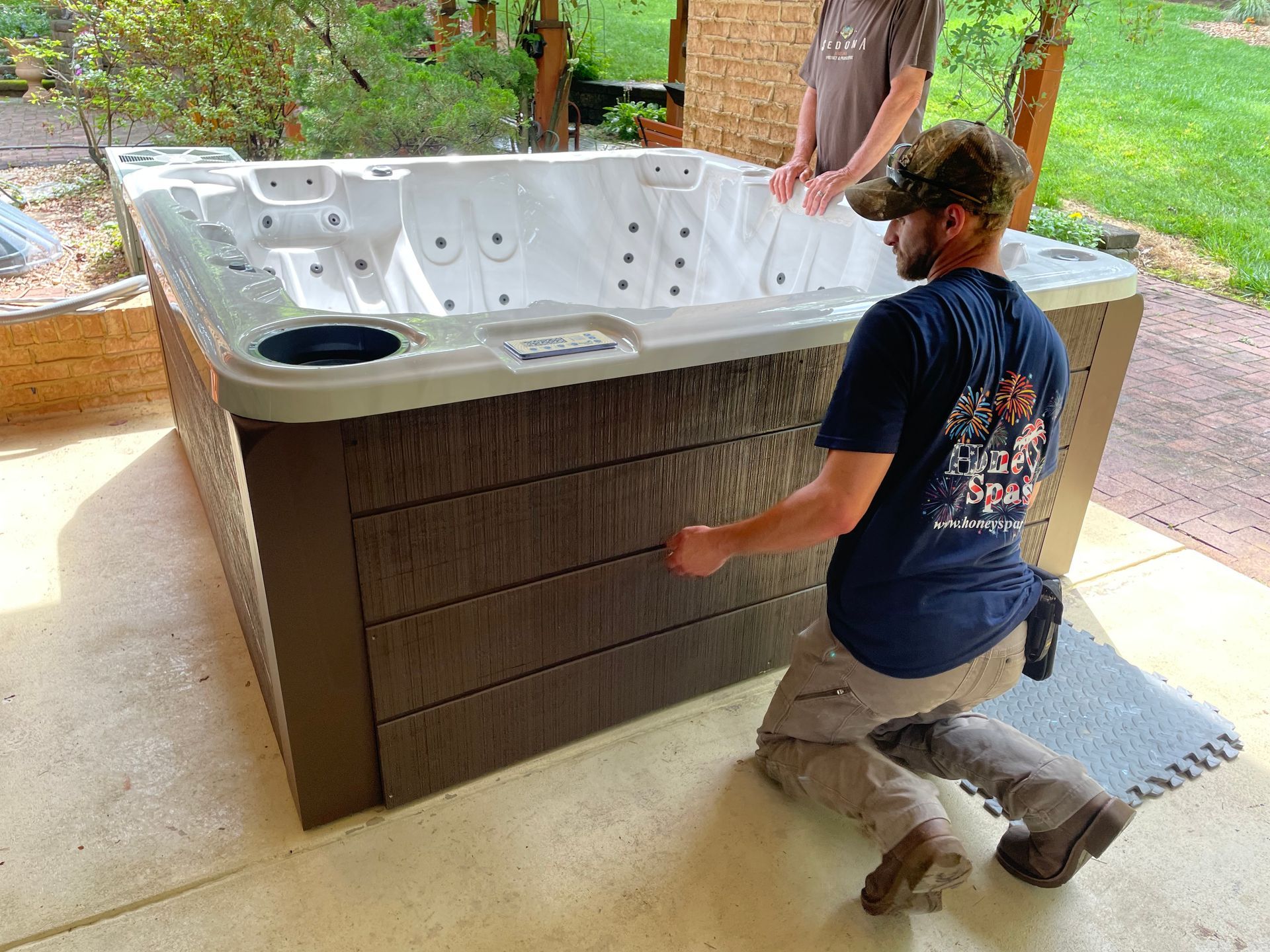 Two men installing a brown-sided hot tub on a concrete patio. One kneels, the other stands.