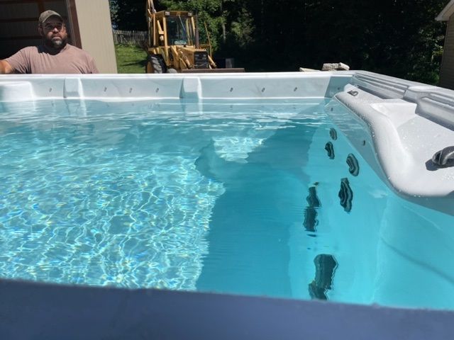 A man looking into a hot tub filled with clear blue water outdoors; a yellow backhoe is in the background.