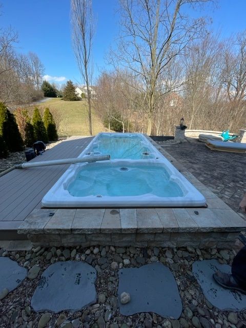 Rectangular swim spa on stone patio with blue water, surrounded by landscaping, clear skies.