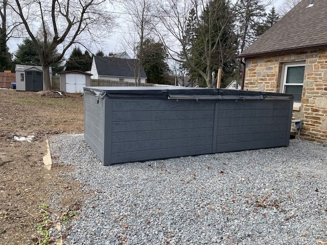 A gray rectangular hot tub on gravel beside a stone building.