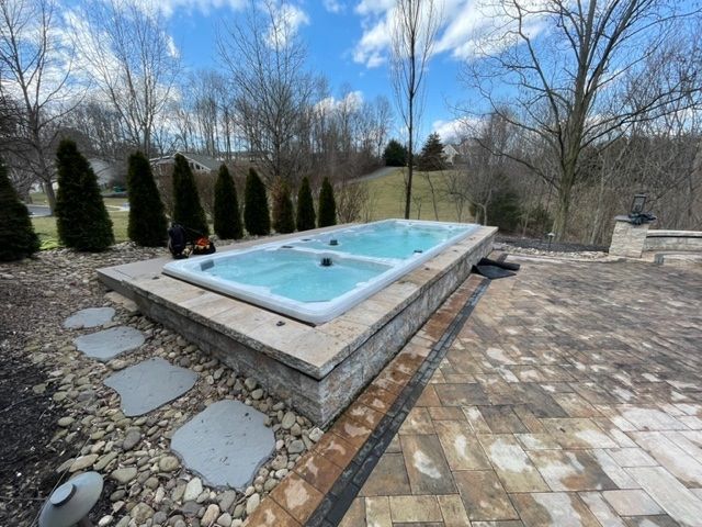 Rectangular outdoor spa with turquoise water, on a stone patio, surrounded by landscaping and trees.