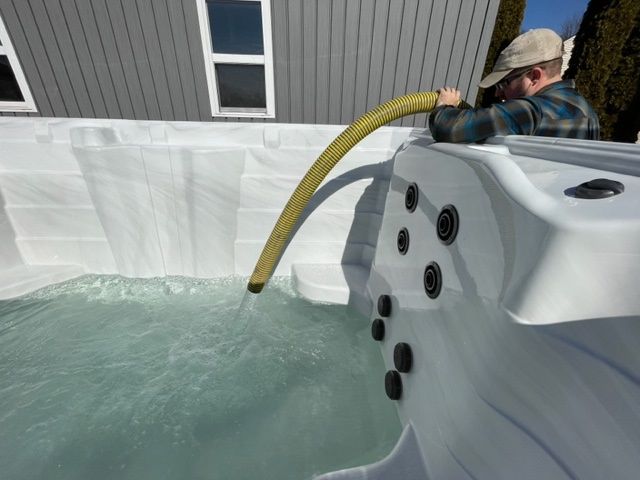 Man filling a white hot tub with a yellow hose. Exterior setting, gray siding in background.