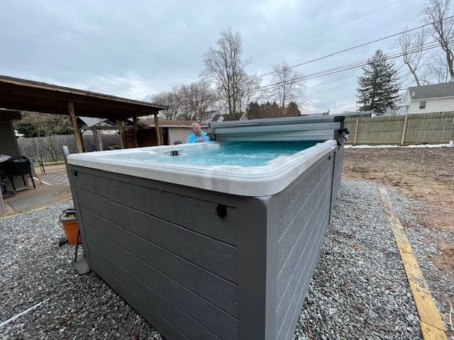 Person in a hot tub with cover open, outdoors. Gray hot tub on a gravel patio with a cloudy sky.