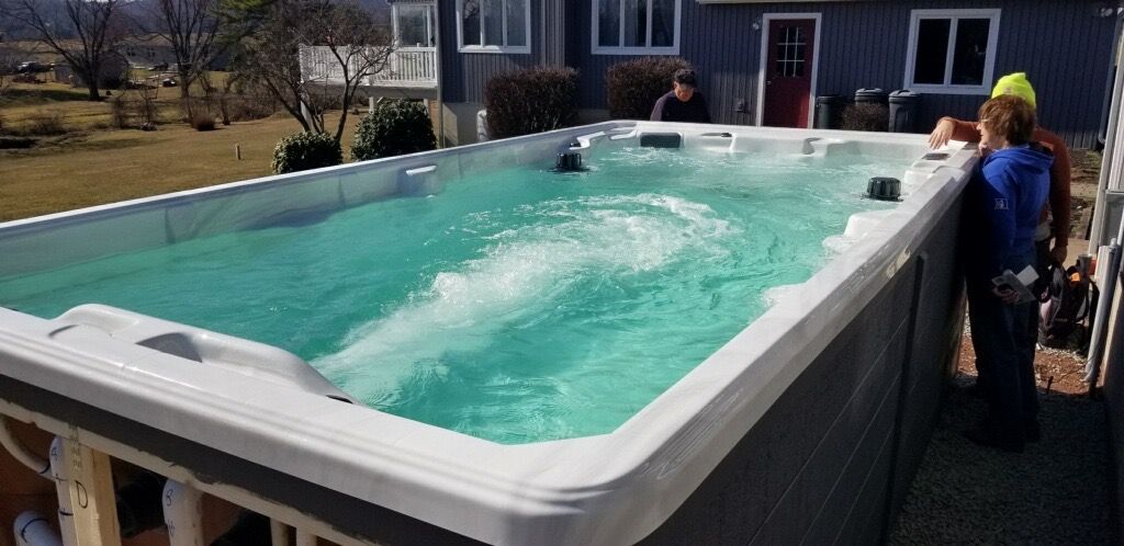 A rectangular swim spa with people standing near it outside a house on a sunny day.
