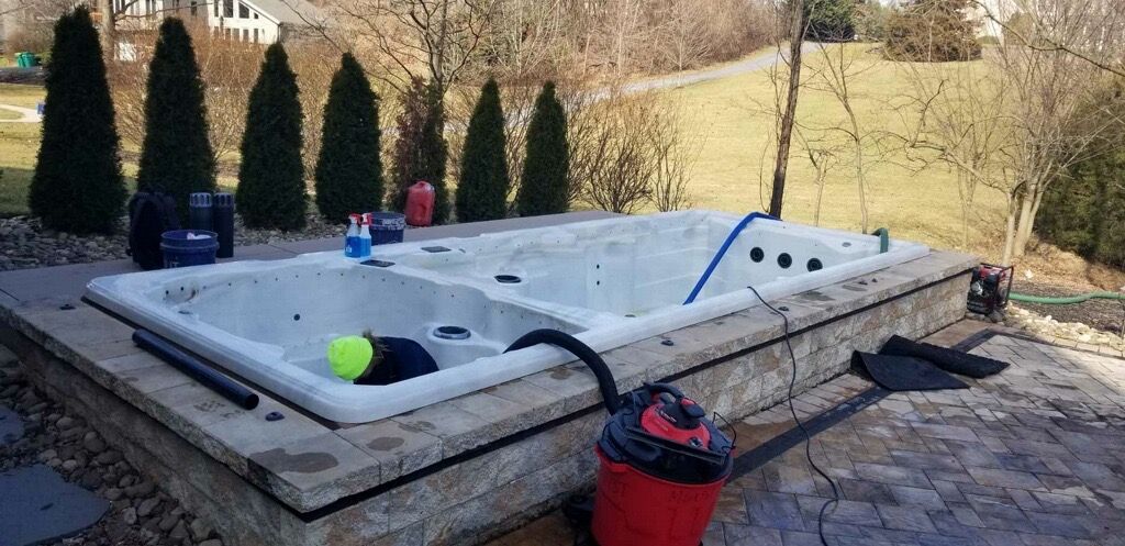 A hot tub sits outdoors, surrounded by a stone patio and trees. A red vacuum is next to it.