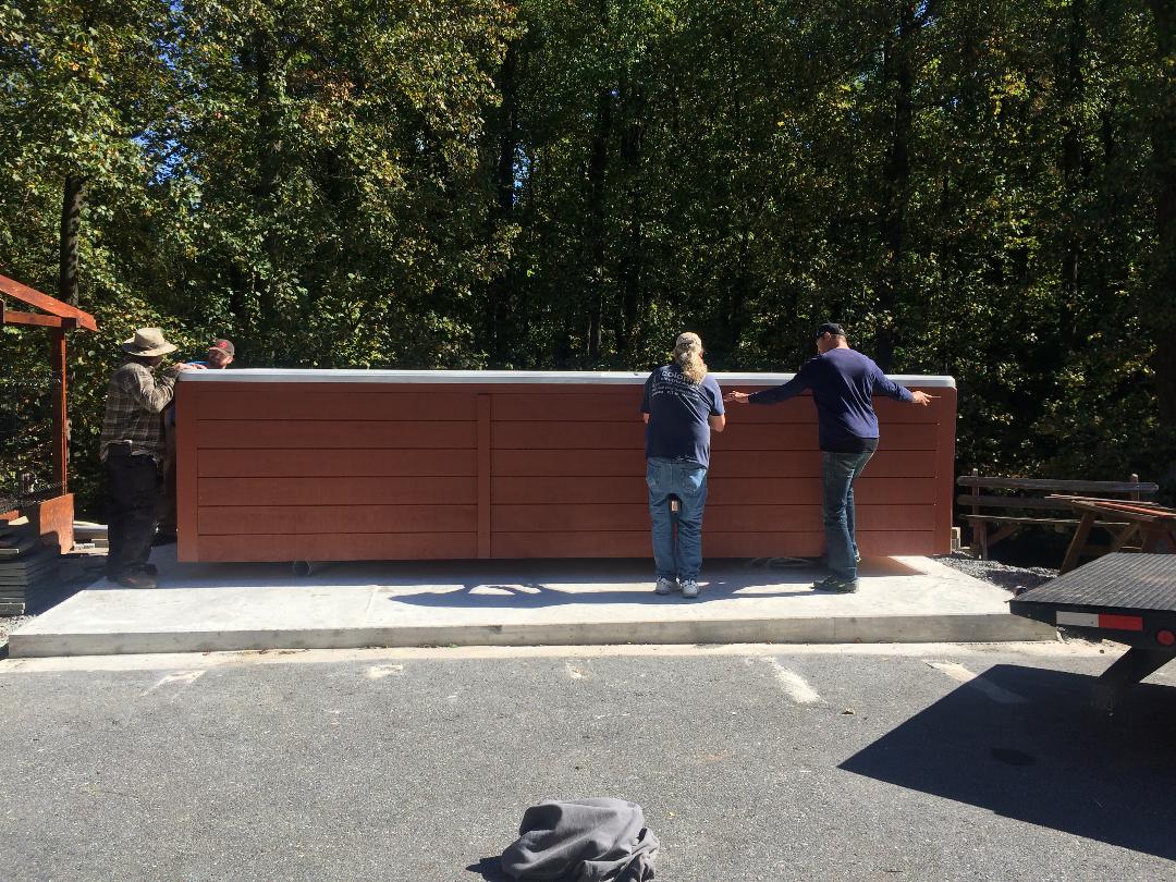 Four people lifting a brown hot tub, outdoors, on a concrete pad.
