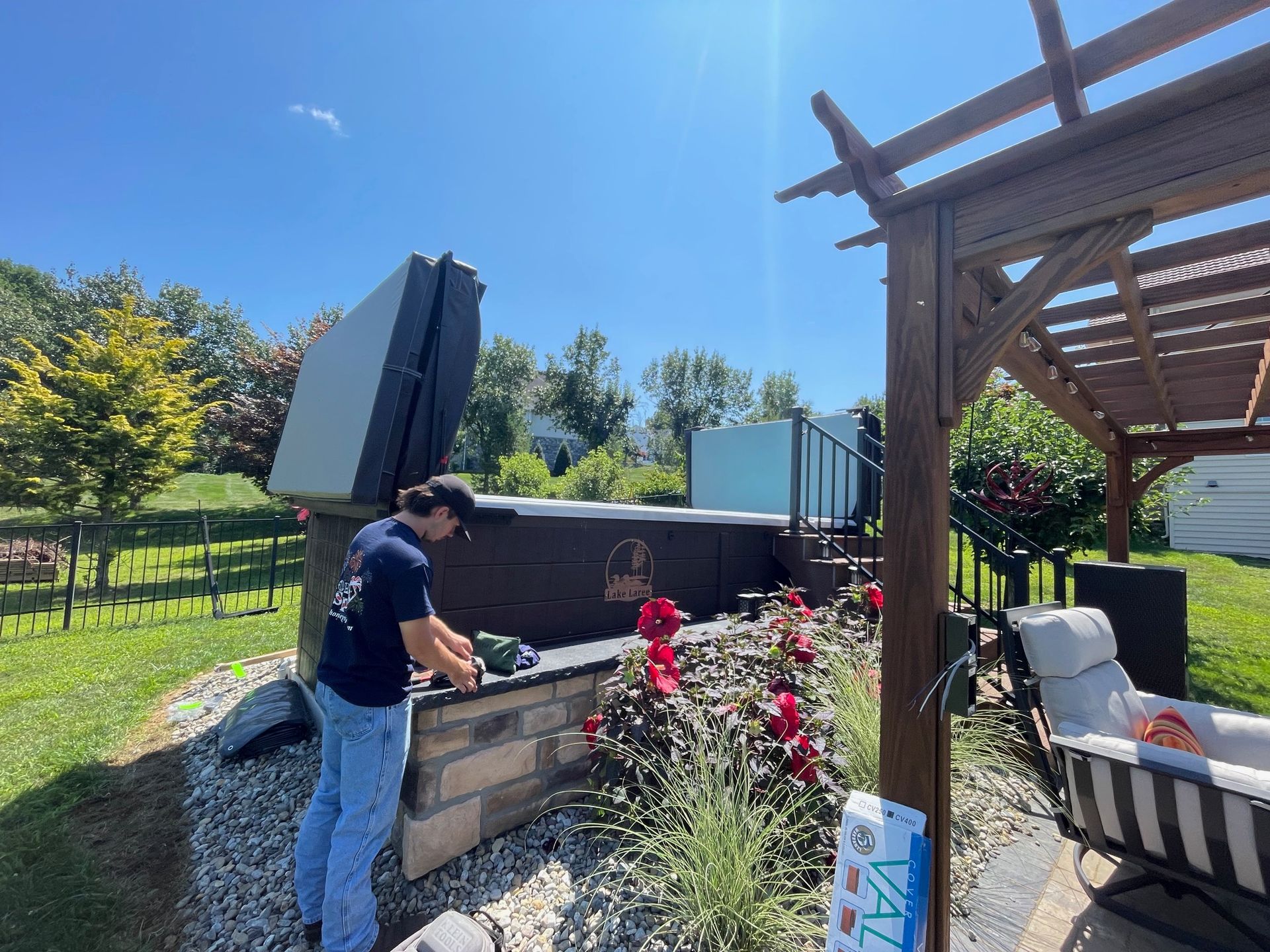 Person working on hot tub outdoors; blue sky, brick and stone surround, pergola, flowers, lawn.
