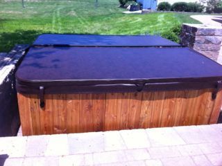 Wooden hot tub with black cover, set on stone patio in a backyard.