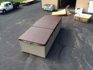 A long, rectangular hot tub with a brown cover on an asphalt lot, next to a building.