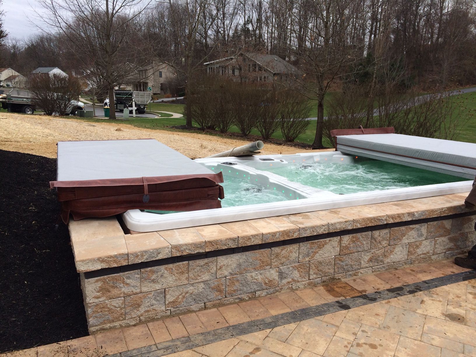 A gray hot tub under a black pergola on a concrete patio, with plants and string lights.