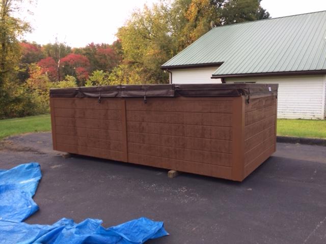 Large brown hot tub with closed cover on wheels, parked on asphalt with a blue tarp.