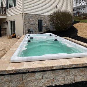 A rectangular swim spa, filled with turquoise water, sits on a stone patio near a house.