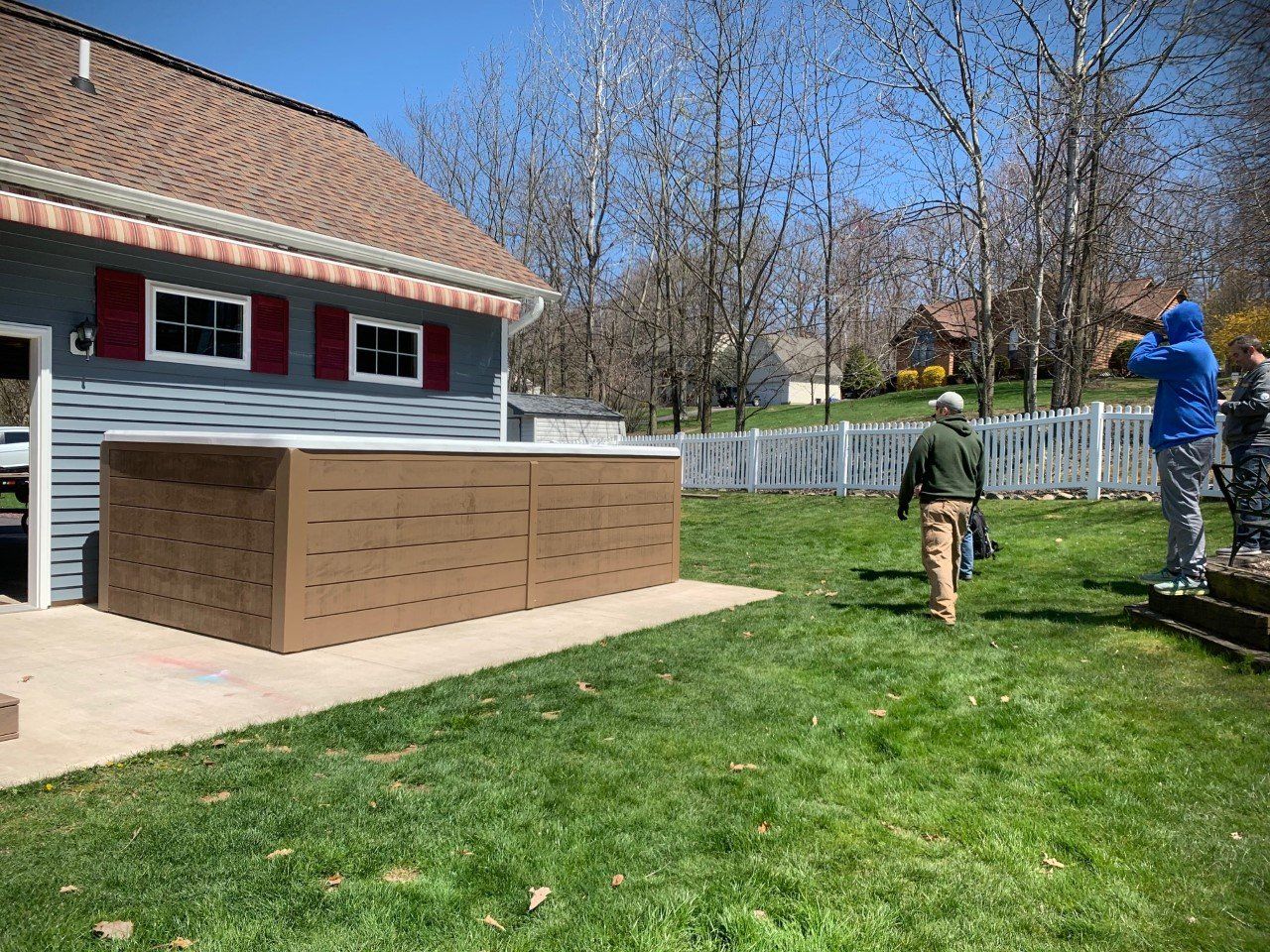 Brown hot tub enclosure near a blue house; two people in the yard, one walking and one standing.
