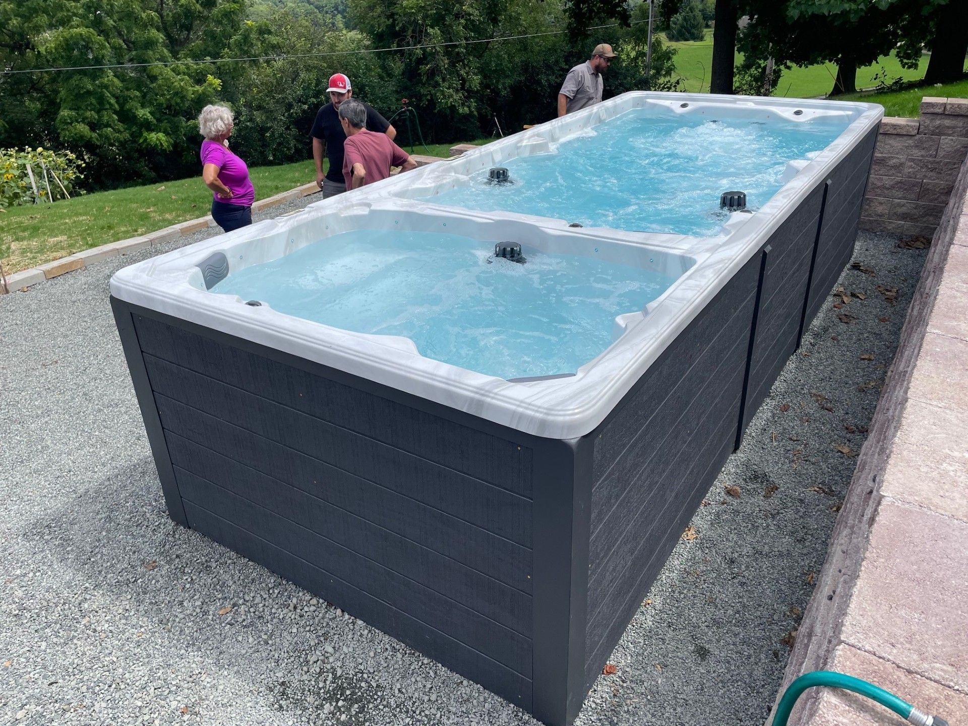 Rectangular swim spa with two sections, filled with water, outdoors. People stand around it.