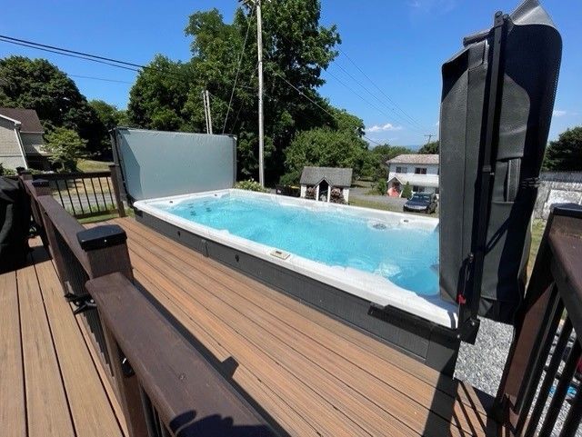 Outdoor swim spa on a wooden deck with open cover, blue water, sunny day.