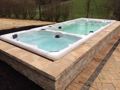A rectangular swim spa with a spa section and a longer swimming area on a stone retaining wall outdoors.