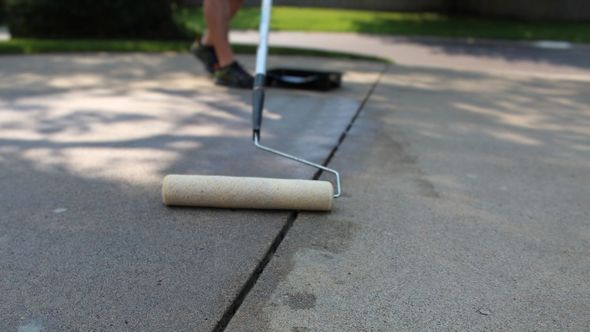 Person using a roller to apply a sealant to a crack in a concrete driveway. Sunlight and trees in the background.