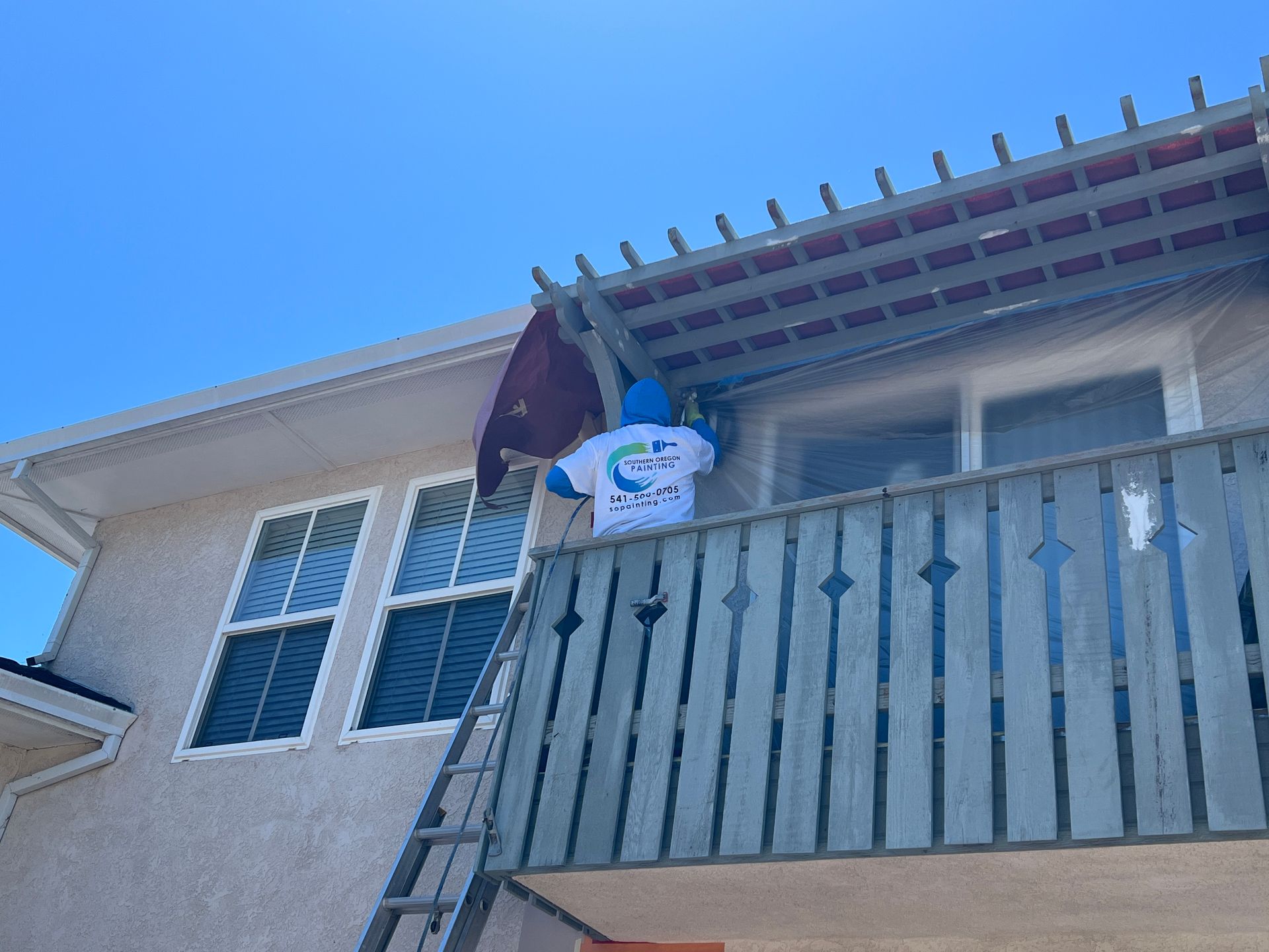 A man is standing on a balcony of a house painting a pergola.