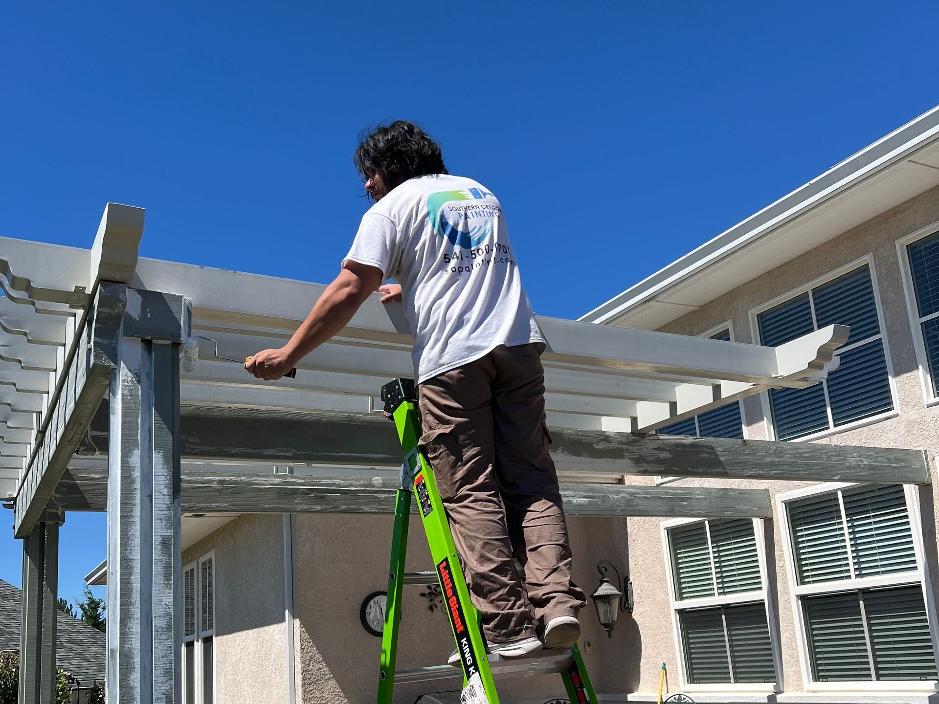 A man wearing a Southern Oregon Painting shirt is standing on a green ladder, working on painting a pergola.