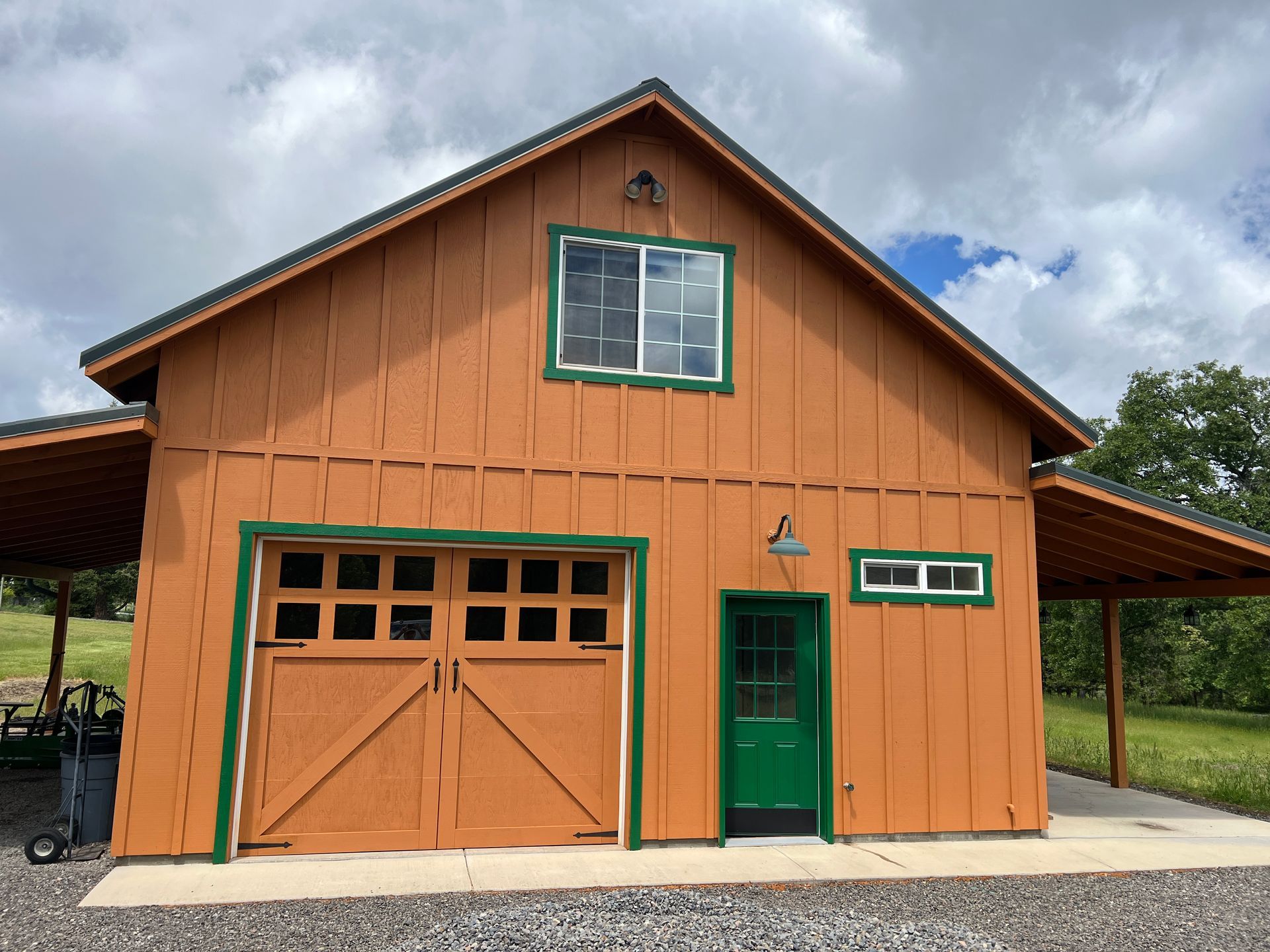 Orange barn with green trim, garage door, and door. White window.  Brown roof extending on either side.