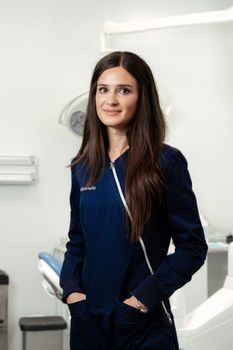 Una donna con una camicetta blu scuro è in piedi in uno studio dentistico, accanto a una poltrona odontoiatrica e alle relative attrezzature.