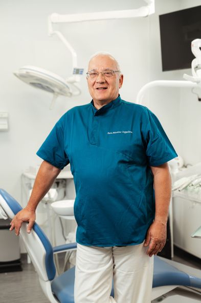Un uomo in piedi in uno studio dentistico, accanto a una poltrona, indossa una casacca color verde acqua e pantaloni bianchi e sorride alla telecamera.