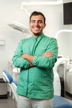 Dentista sorridente in camice verde, in piedi con le braccia incrociate in uno studio dentistico.