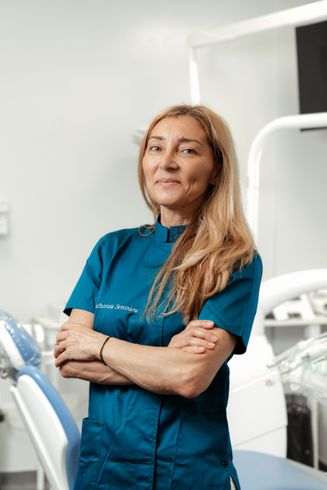 Un'operatrice sanitaria in camice color verde acqua è in piedi con le braccia incrociate in una stanza clinica e sorride alla telecamera.