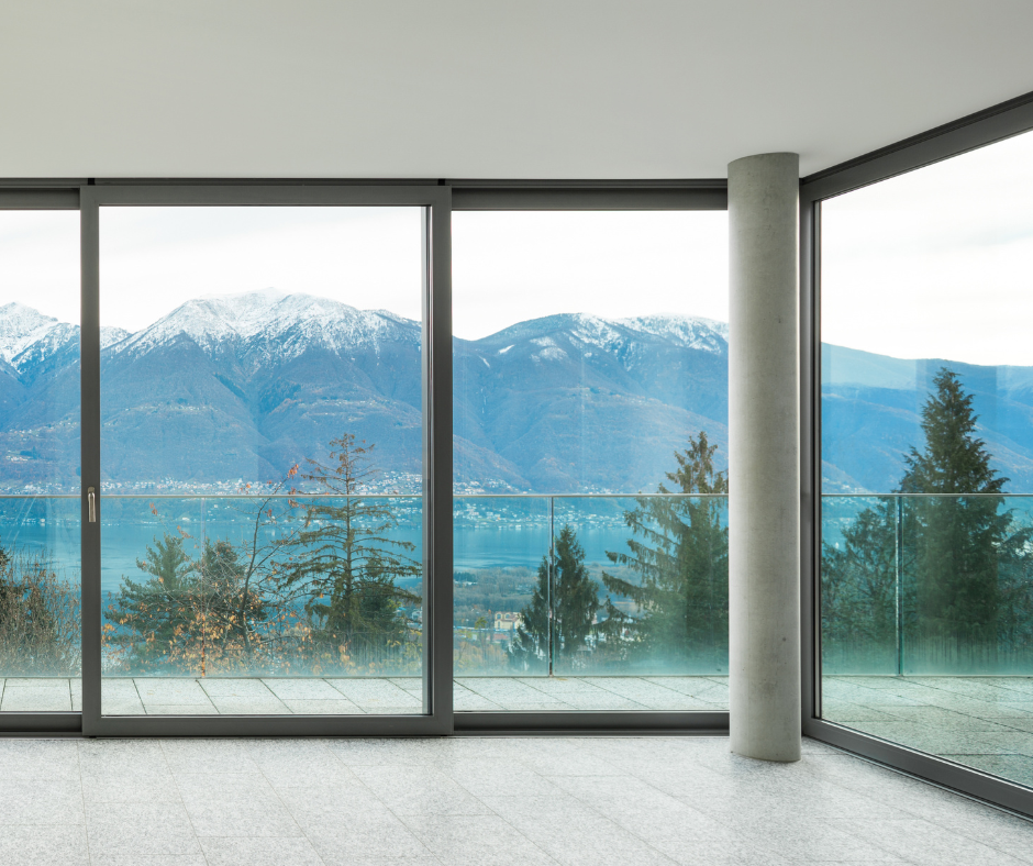 Chambre avec de grandes fenêtres donnant sur un lac et des montagnes enneigées. Encadrements gris, sol en pierre et pilier.
