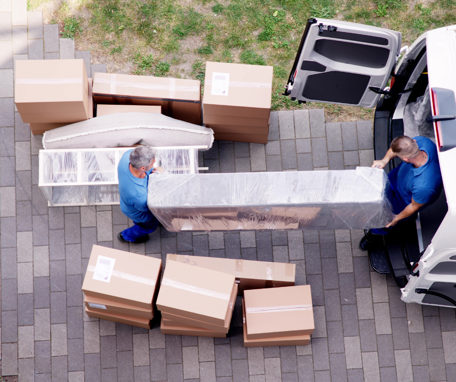 Des déménageurs chargent un objet volumineux et emballé dans une camionnette, entourée de cartons sur une allée pavée.