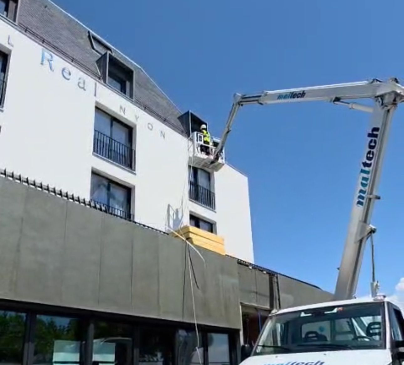 Un ouvrier du bâtiment répare la façade d'un immeuble depuis une nacelle. L'enseigne « Hôtel Rea NYON » est visible. Ciel bleu dégagé.