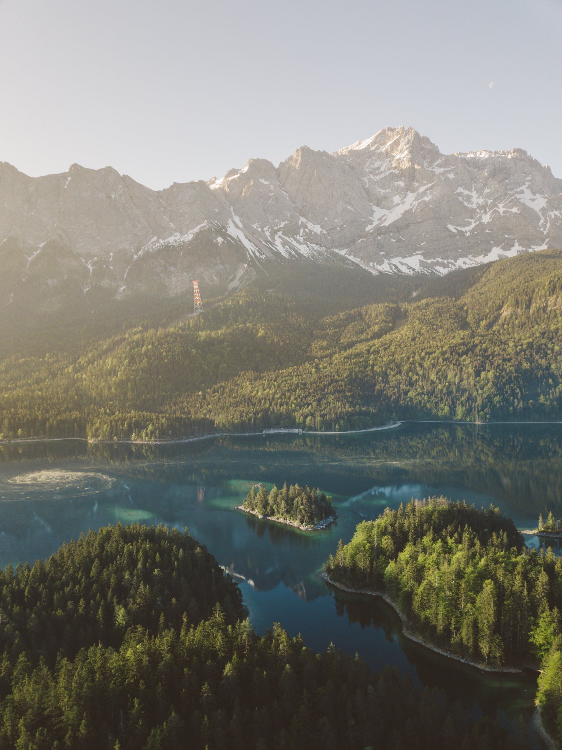 Üppiger grüner Wald umgibt einen klaren blauen See mit Inseln, darunter schneebedeckte Berge im Sonnenlicht.