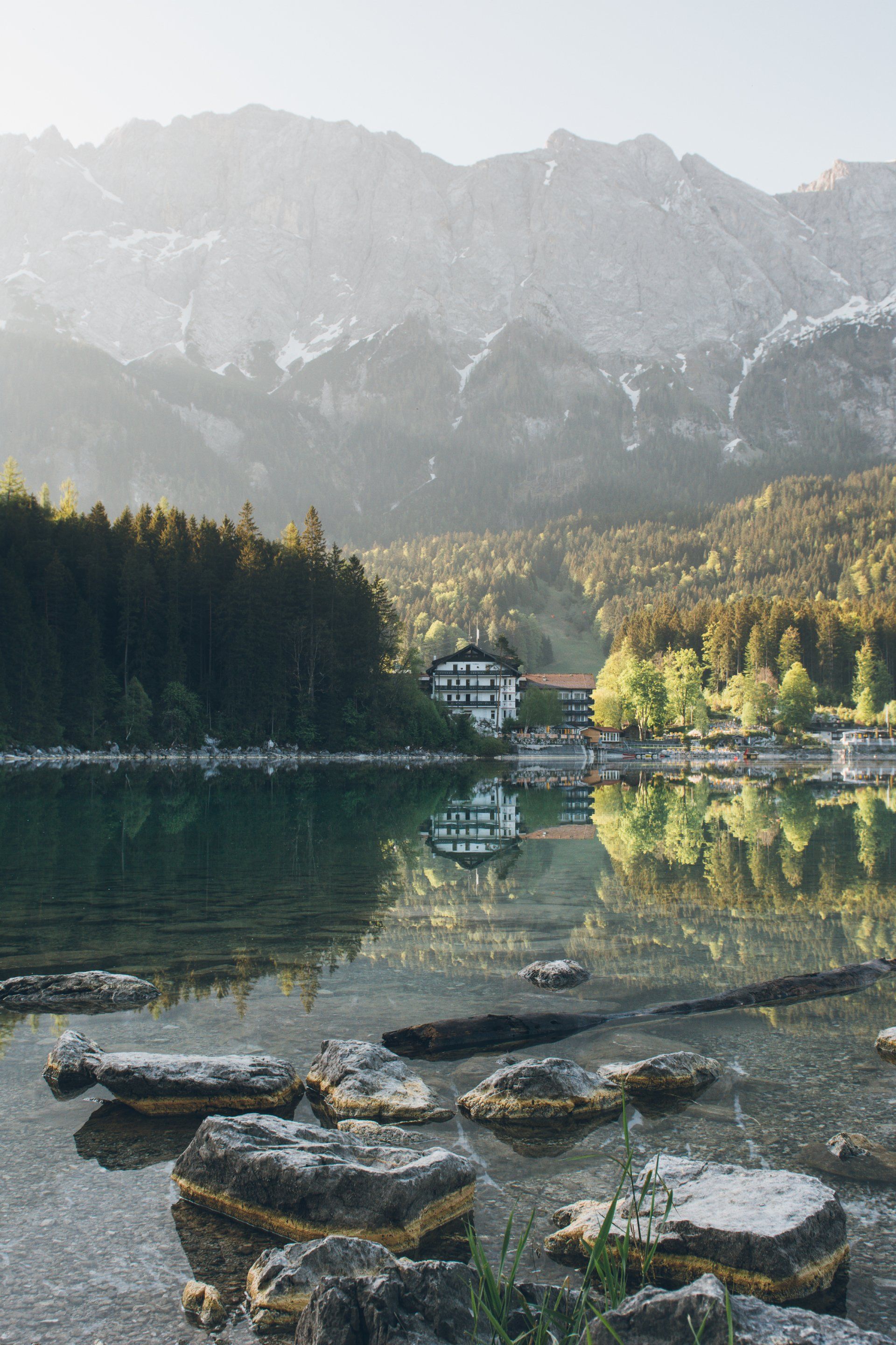 Eibsee mit felsigem Ufer, Bäume, Gebäude und Berge spiegeln sich im Sonnenlicht.