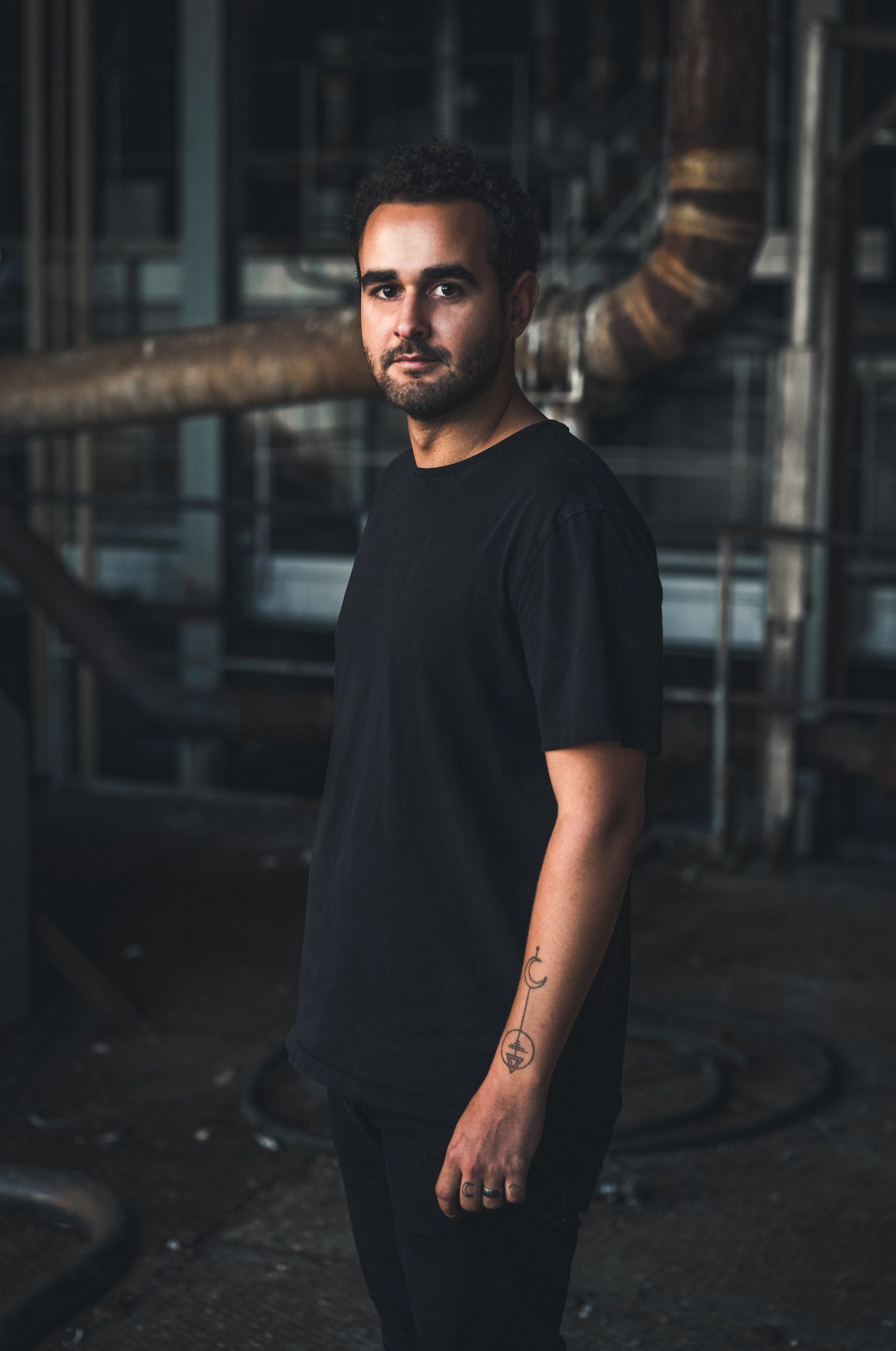 Mann in schwarzem Hemd und Hose steht in Industrieumgebung und blickt in die Kamera.