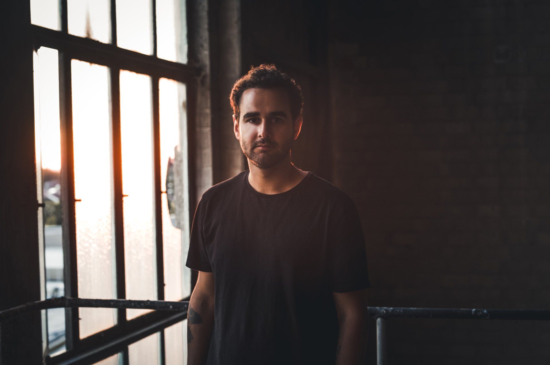 Ein Mann steht im Sonnenlicht an einem Fenster. Er trägt ein schwarzes Hemd und blickt in die Kamera.