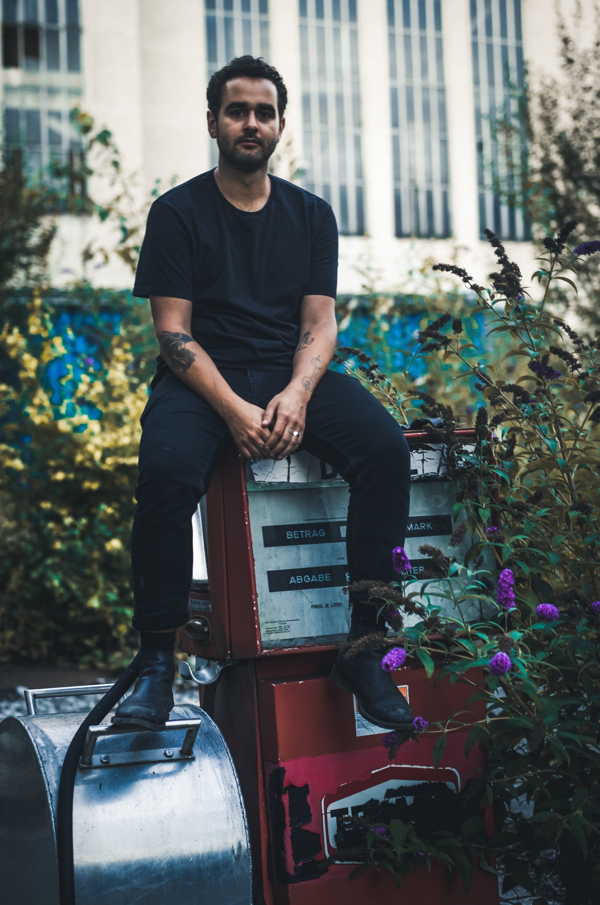 Mann in schwarzer Kleidung sitzt auf einer rostigen Zapfsäule, Pflanzen im Hintergrund.