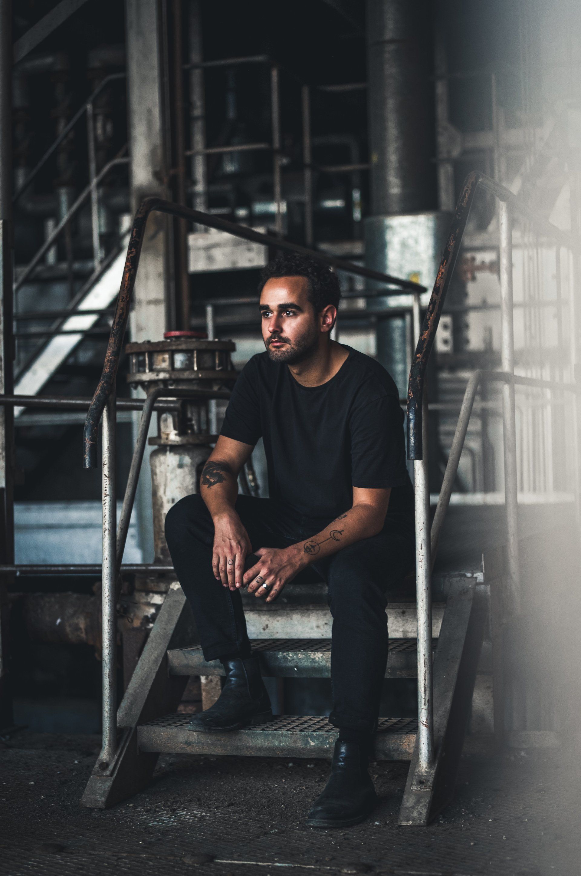 Ein Mann in schwarzer Kleidung sitzt nachdenklich auf einer Metalltreppe in einem Industriegebiet.