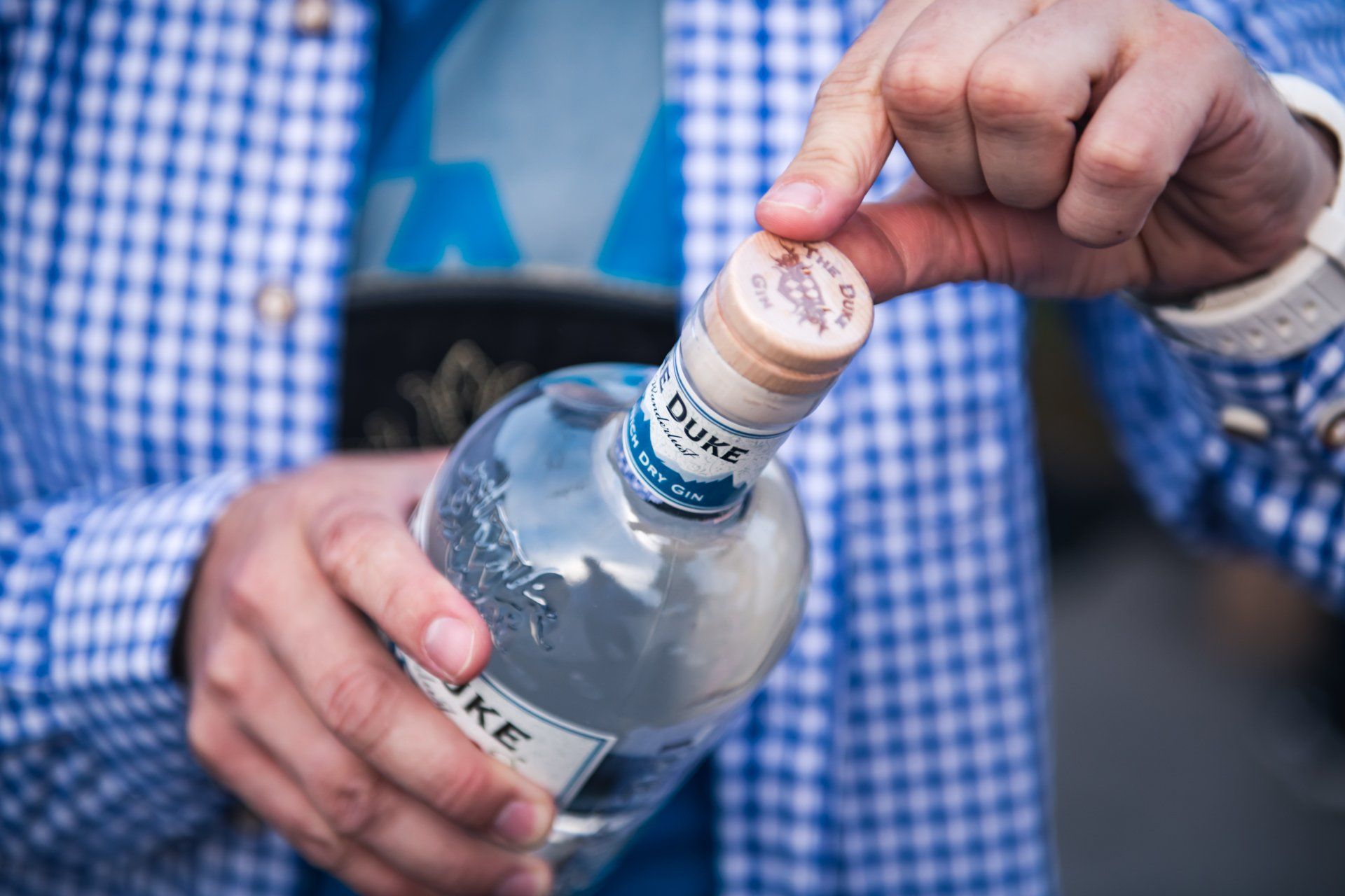 Person, die eine Flasche Duke Gin öffnet und eine blau karierte Jacke trägt.