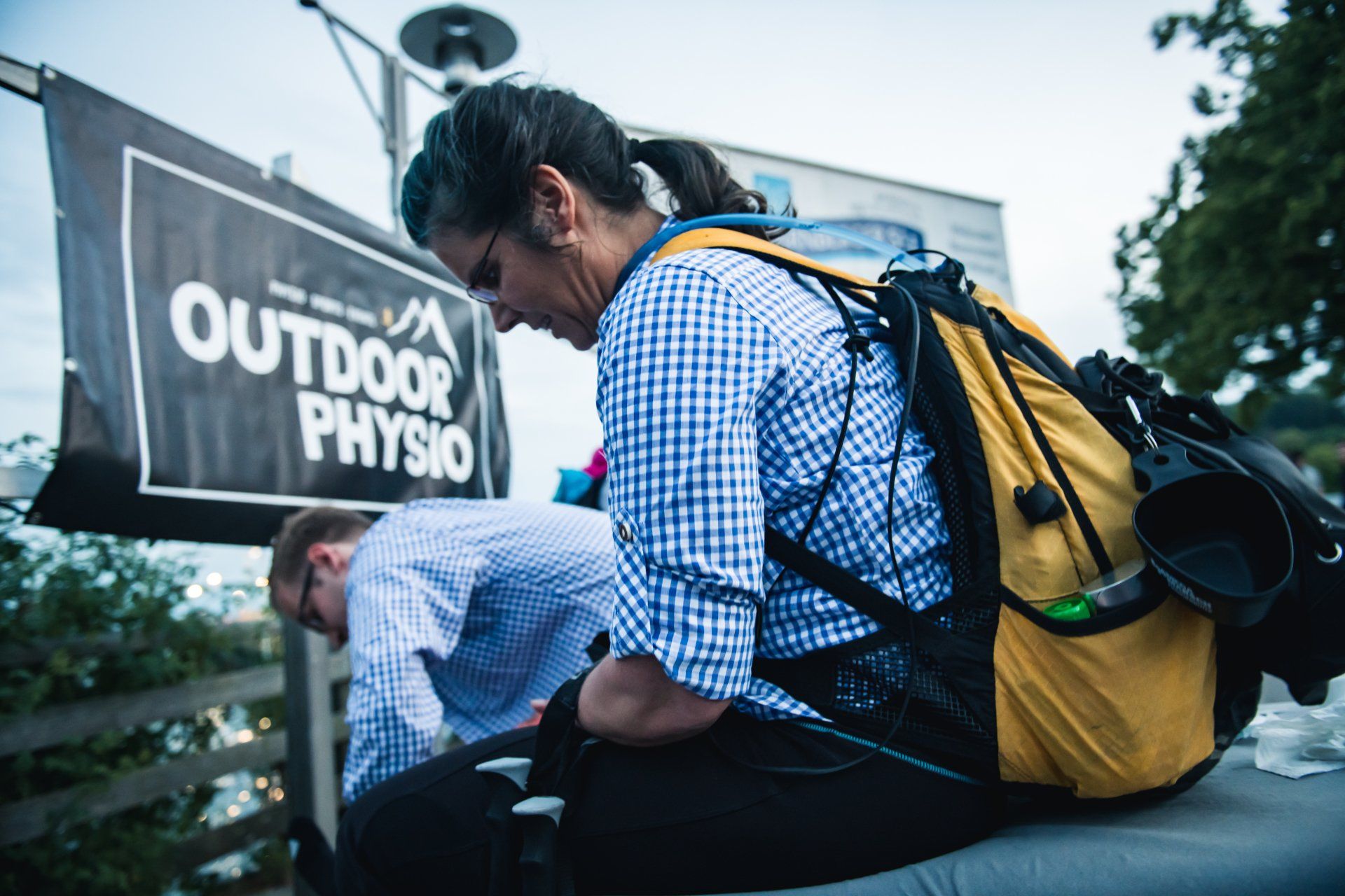 Frau mit Rucksack lehnt sich im Freien nach unten, neben einem Schild mit der Aufschrift „Outdoor Physio“.