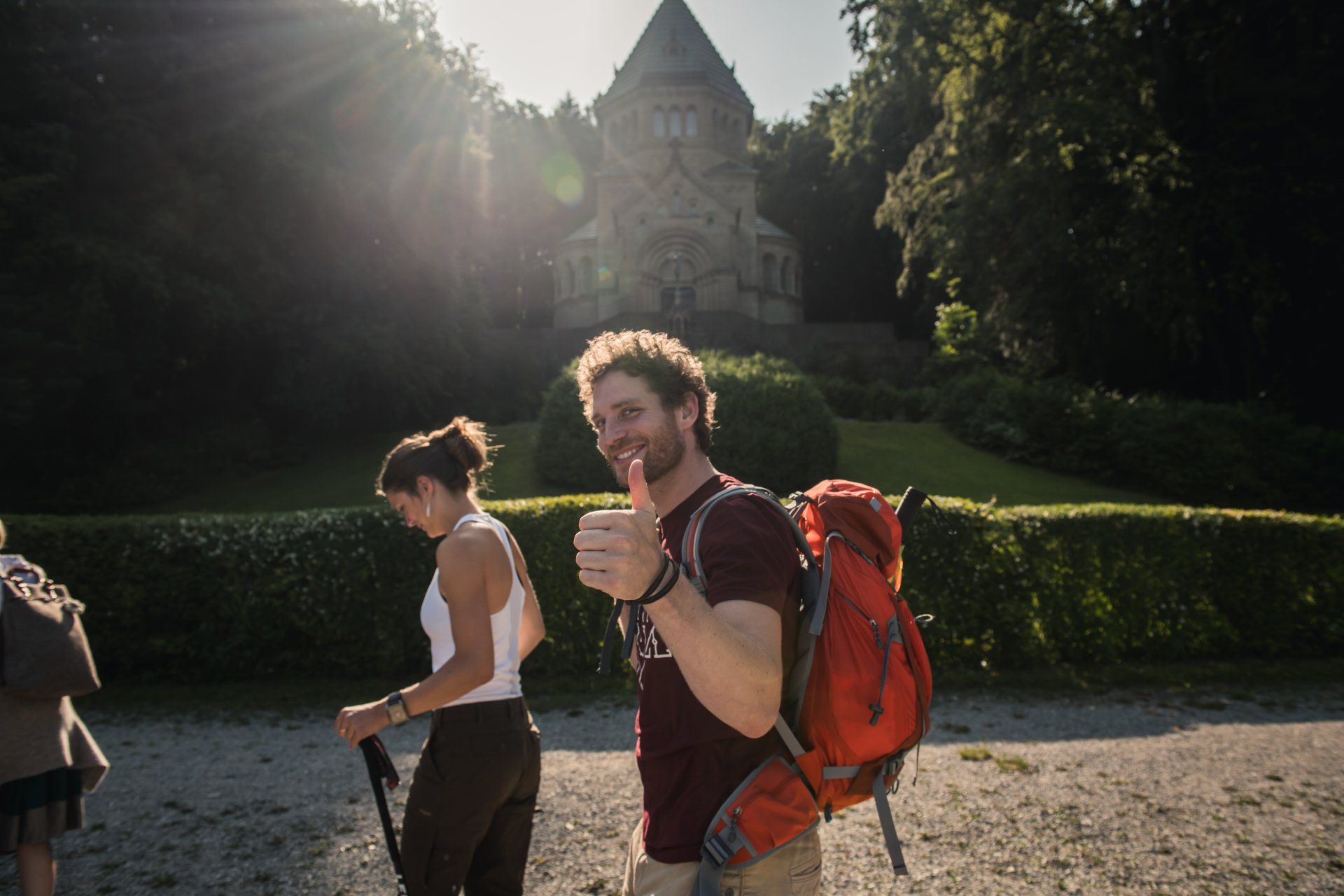 Mann mit Rucksack zeigt Daumen hoch und wandert mit anderen in der Nähe eines Steinturms in sonnenbeschienenen Wäldern.
