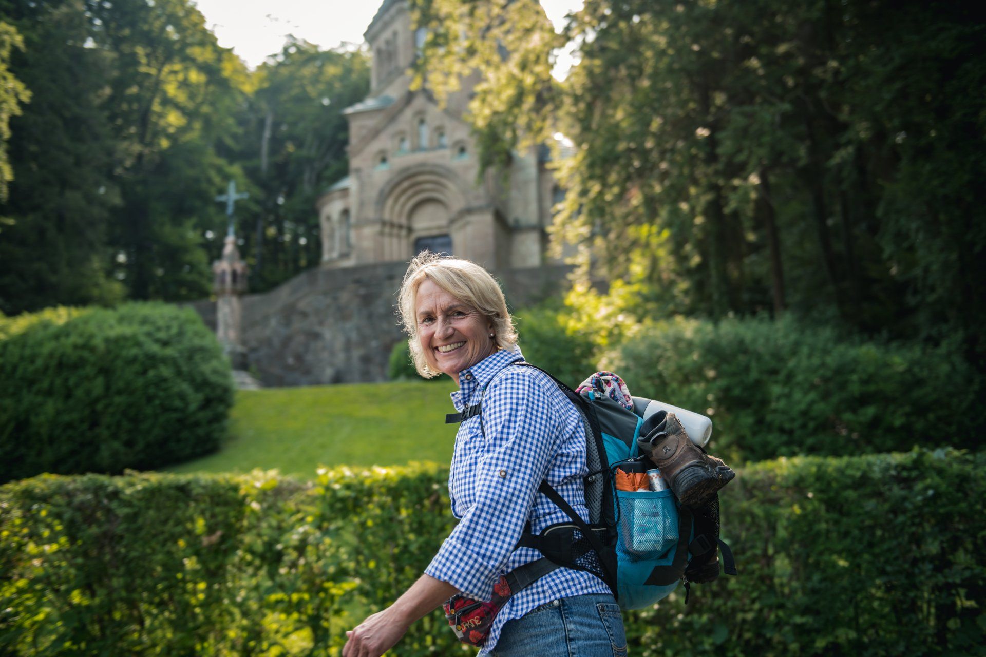 Frau mit Rucksack lächelt beim Wandern in der Nähe eines Steingebäudes mit einem Kreuz in einer grünen, sonnigen Umgebung.